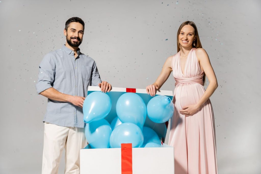 Positive pregnant woman in elegant dress opening bog gift box with blue balloons while standing near husband under confetti during baby shower on grey background, gender party, it`s a boy