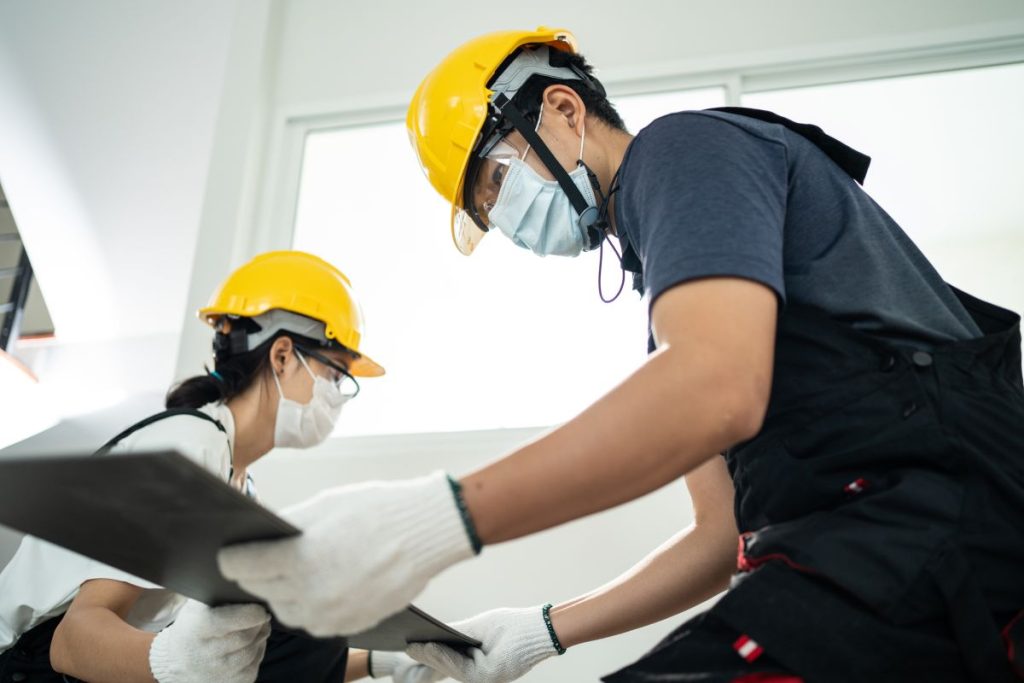 Asian Craftsman or Carpenter worker man and woman wear safety helmet and protective face mask due to Covid-19 pandemic. Joiner Builder installs laminate board on floor to renovating apartment or house