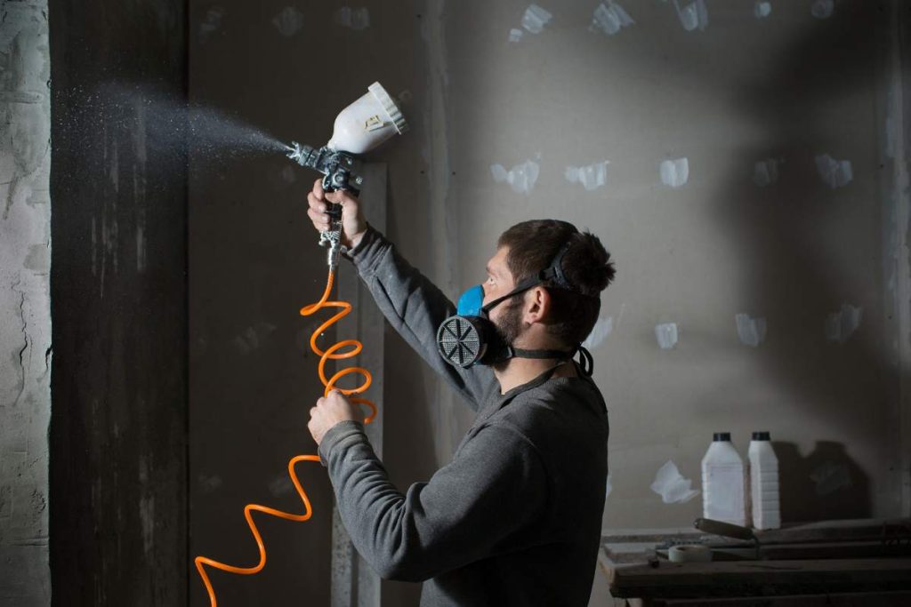 a male plasterer in a construction respirator paints a wall from a spray gun before plastering and putty.