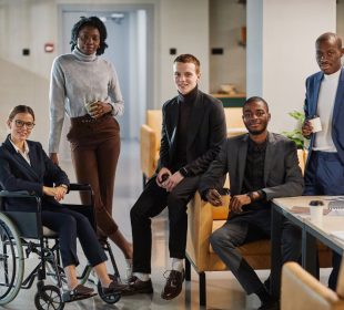 Full length portrait of diverse business team with young woman in wheelchair all smiling at camera in office