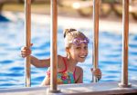 A little girl in a bright pink swimsuit with transparent black goggles for swimming, straightens her goggles and then dives into a deep blue pool with a chimta of clear water