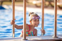 A little girl in a bright pink swimsuit with transparent black goggles for swimming, straightens her goggles and then dives into a deep blue pool with a chimta of clear water