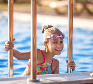 A little girl in a bright pink swimsuit with transparent black goggles for swimming, straightens her goggles and then dives into a deep blue pool with a chimta of clear water