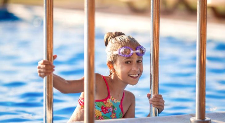 A little girl in a bright pink swimsuit with transparent black goggles for swimming, straightens her goggles and then dives into a deep blue pool with a chimta of clear water