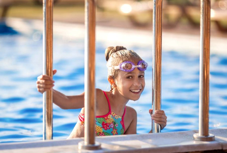 A little girl in a bright pink swimsuit with transparent black goggles for swimming, straightens her goggles and then dives into a deep blue pool with a chimta of clear water