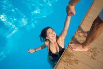 View from above of attractive girl in the swimming pool holding a man's hand trying to get out at resort. Sunny day